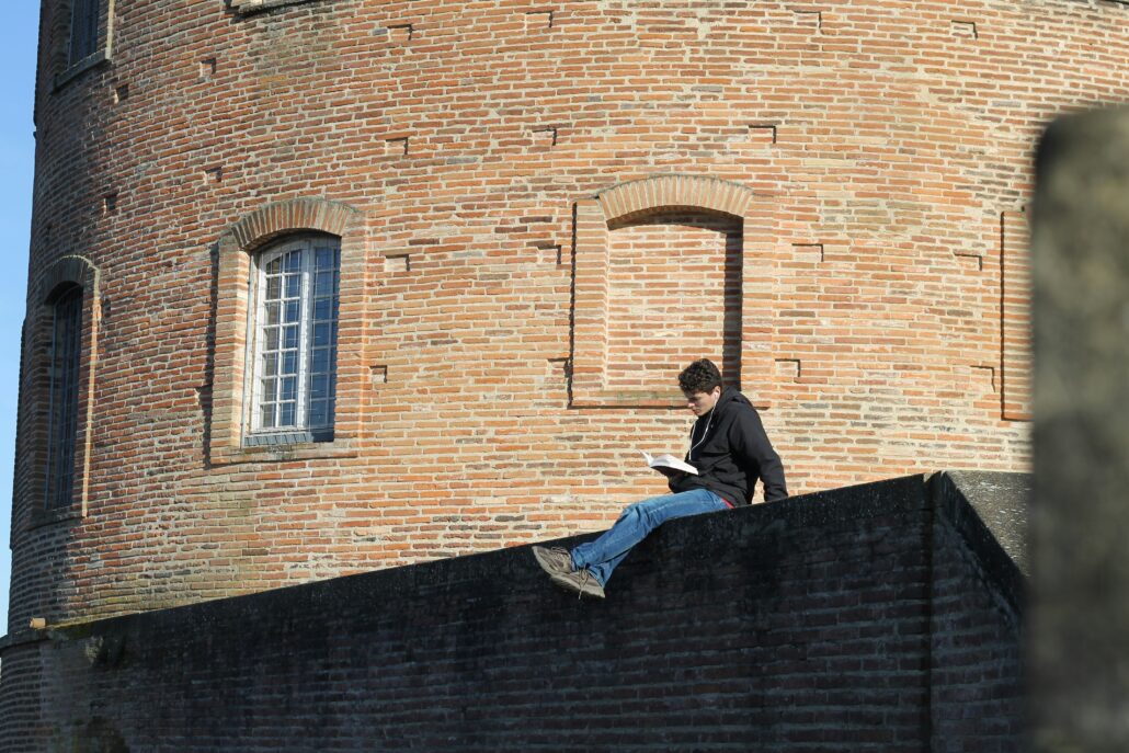 Une jeune homme assis devant un bâtiment en briques rouge lit un livre.
